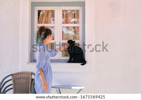 Teenager girl touching beautiful black cat sitting on the window sill outdoor, sunny summer day, copy space white wall background