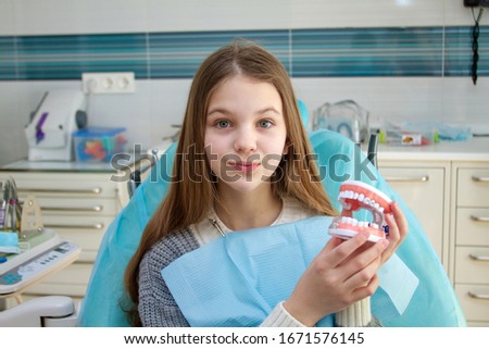Female teenage girl patient holds 3d model of fake teeth with braces looking at camera. Concept of bite correction appliance demonstration for medical cosmetic purposes. Dental clinics interior.
