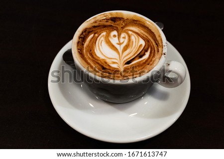 A cup of cappuccino with a flower in the milk foam on black background - latte art coffee shop concept