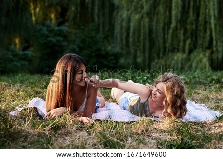 Two sisters, lying on blanket on grass in park in summer. Pretty girls, wearing light blue jeans shorts and green beige top, relaxing outside, tickling each other. Summertime leisure outside.