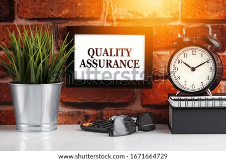 QUALITY ASSURANCE text with alarm clock, books and vase on brick background. Business, Quotes and Copy Space concept