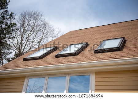 triple skylights on red roof with yellow siding and blue sky with trees in background Royalty-Free Stock Photo #1671684694