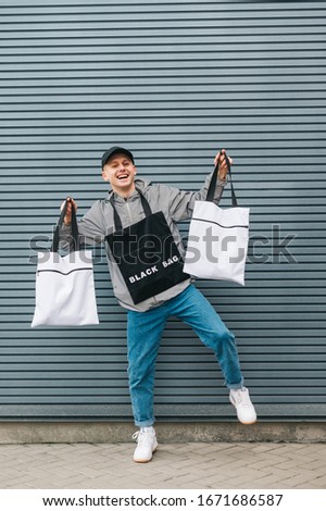 Full length photo of eco friendly young man in stylish streetwear standing on a gray background with eco bags in his hands and posing at camera with a smile on his face. Vertical. Copy space