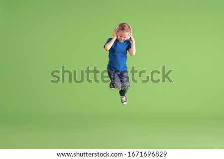 Listen to music. Happy boy playing and having fun on green studio background. Caucasian kid in bright looks playful, laughting, smiling. Concept of education, childhood, emotions, facial expression.