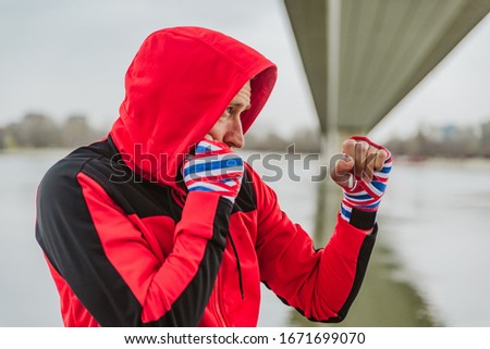 A boxer with a bandage on his arms, in a red hooded sweatshirt, is training outdoors
