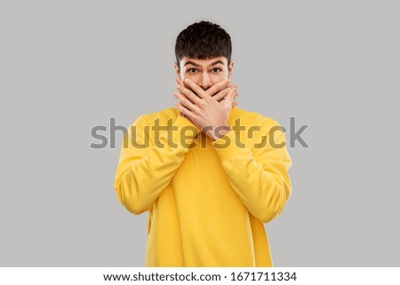 emotion, expression and people concept - shocked and speechless young man in yellow sweatshirt covering his mouth with hands over grey background