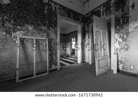 A black and white photograph inside an abandoned house with an open doorway, taken in the ghost town of Kolmanskop, Namibia.