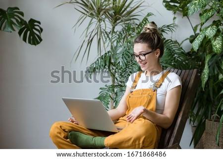 Woman gardener in glasses wearing overalls, sitting on wooden chair in greenhouse, using laptop after work, talks with her friend about coronavirus and stay home during online video call. Working home Royalty-Free Stock Photo #1671868486