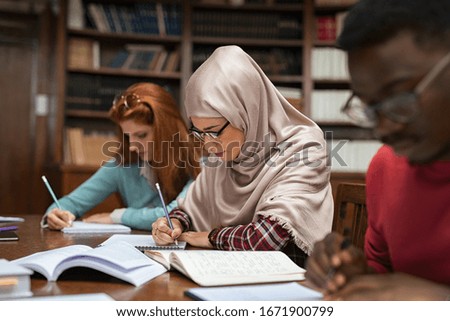 Focused student in hijab studying with classmates in university library. Islamic young woman in spectacles wearing abaya and writing in notebook. Arabian girl studying with multiethnic students.