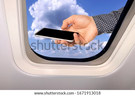 male hand with a smartphone on the background of a beautiful sky blue sky landscape with fluffy clouds behind the porthole of a plane, travel concept