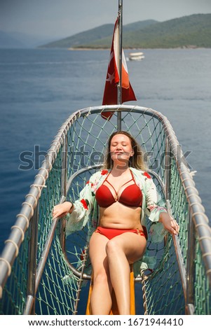 Curvy woman in a red swimsuit sitting on the front of the boat deck on turqouise blue sea background. View from the back - Sea voyage or cruise concept.