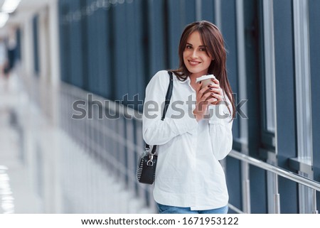 Brunette in white shirt indoors in modern airport or hallway at daytime and holding cup of drink.
