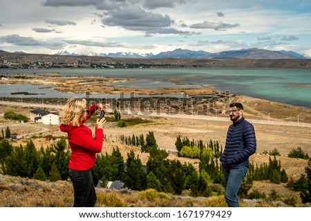 A young woman takes a picture with her boyfriend's phone against the backdrop of a beautiful lake and landscape in patagonia.