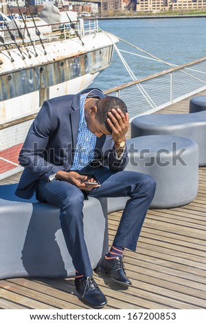 Dressing in a blue suit, leather shoes, a hand touching the top of his head , a young black businessman is sitting on the deck, looking down at his mobile phone / Waiting for Phone Call