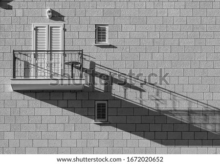 Black and white image of a single modern balcony on paving stone wall, white shutters door, shadow on the wall