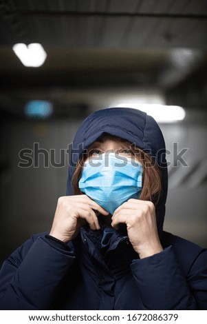 White woman with brown hair wearing a dark coat with a hood and a blue face mask against virus infections is standing in an underground parking lot
