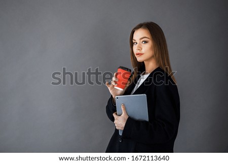 Young pretty girl in business style clothes stands on a gray background with a tablet and a cup of coffee in her hands