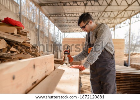 Young man carpenter at the wooden workshop warehouse is using a milling machine on a wooden plank/board from a big wood timber stack of wooden planks.