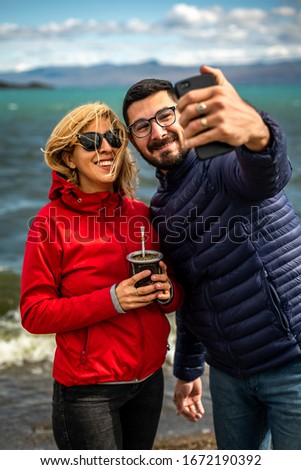 Happy couple taking a selfie with a phone camera on the background of a beautiful lake with blue water and drink yerba mate. Spending time together, travel, happiness concept