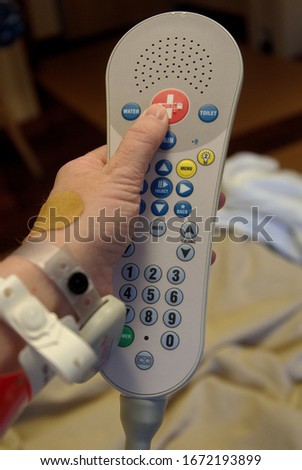 Pressing the "Call for Nurse" red button on a remote, with patient bed in background, at a local hospital