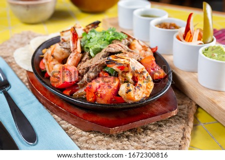 A view of a sizzling skillet plate of surf and turf style fajitas, featuring several condiments and dips on the side.