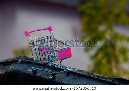 Photograph of a toy luggage trolley made of metal and plastic. the trolley has pink colour with out of focus trees in the background