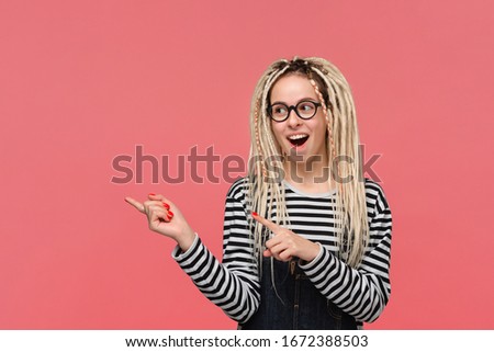 Beautiful young girl with dreadlocks in a striped shirt and jeans jumpsuit pointing with two fingers at copy space. Place for advertising