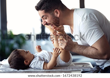 Shot of smiling young father has fun with little baby while changing his nappy at home. Royalty-Free Stock Photo #1672438192