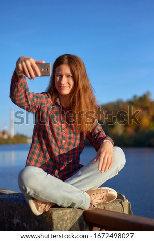 Happy young woman in plaid shirt taking selfie sitting on iron railing at river embankment in sunny autumn day. Lifestyle relax recreation weekend concept