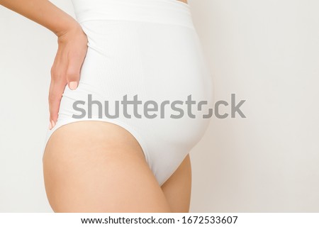 
Young woman in white long, large panties on light gray background. Underwear before and after pregnancy time. Closeup. Side view.