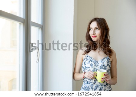 Brunette girl with long hair looks out the window with a yellow cup of tea. She looks out.