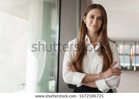 Pleasant young female assistant, manager standing in office corridor near window, with pleased confident expression, cross hands over chest showing readiness, business and women concept Royalty-Free Stock Photo #1672586725