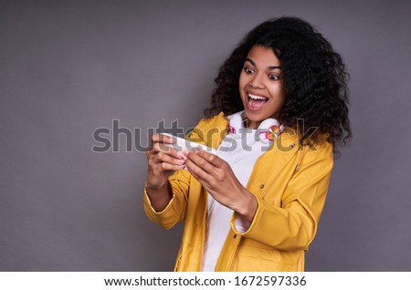 Reckless cheerful joyful playful young African girl, in a white T-shirt, with curly curvy hair, holds a smartphone in her hand, plays online games, a young gamer, plays on a gray background.