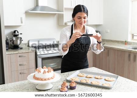 Gorgeous female blogger smiling while taking pictures of cookies from smartphone for social media in kitchen