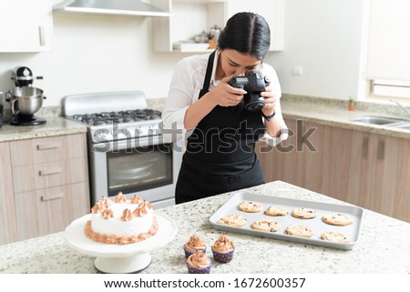 Young female food photographer taking pictures of fresh cookies in tray by cake and cupcakes on counter at home