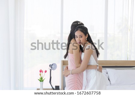 An Asian bride in a white wedding dress embraces her friend in a pink dress on her wedding day with a feeling of gratitude.