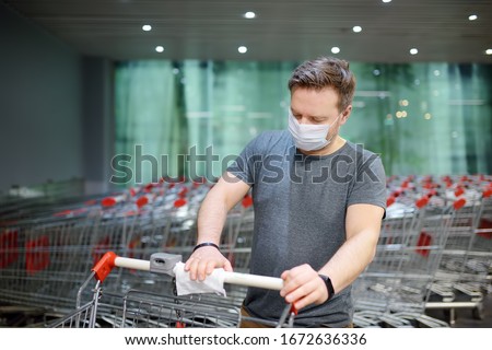 Man wearing disposable medical face mask wipes the shopping cart handle with a disinfecting cloth in supermarket. Safety during coronavirus outbreak. Epidemic of virus covid. Royalty-Free Stock Photo #1672636336