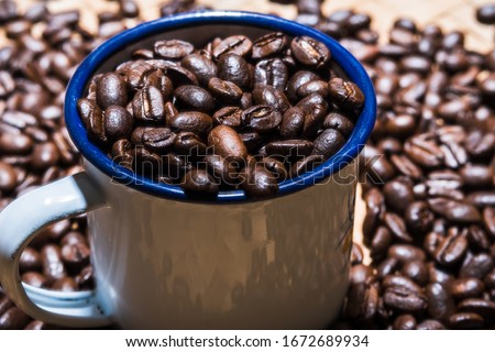 Dark roasted coffee beans in rustic enamel mug, and some others scattered around it as a blurry background.