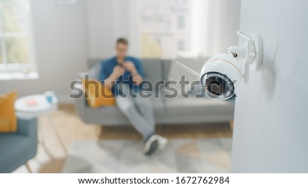 Close Up Object Shot of a Modern Wi-Fi Surveillance Camera with Two Antennas on a White Wall in a Cozy Apartment. Man is Sitting on a Sofa in the Background. Royalty-Free Stock Photo #1672762984