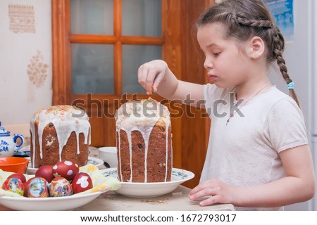 Easter cakes stand on a table in a village house. Girl decorates Easter cakes with caramel. Easter eggs lie in a large dish. Girl 5 years old, European. in home clothes. Photo of a real situation.