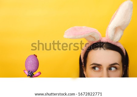 Close up portrait of young woman wears bunny ears, hides face, looks mysteriously from underneath on a purple Easter egg, isolated on yellow background. Spring holiday and Easter concept.
