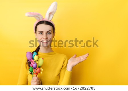 Studio shot of pleased woman wears bunny ears, raises palm as if showing something, gestures over blank space, holds colored eggs, dressed in sweater, models over yellow wall. Seasonal holiday concept