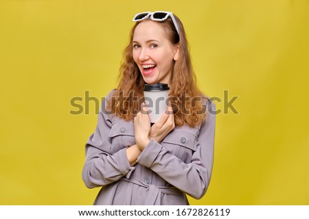 Stylish caucasian girl in a gray dress on a yellow background. Holding a disposable coffee cup to go with both hands. Copy space. Close up.