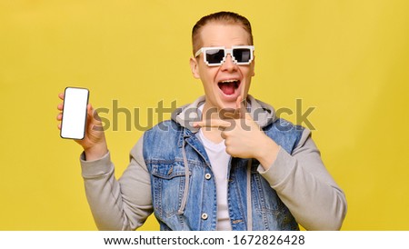 Stylish Caucasian man in a jeans on a yellow background with 8 bit glasses. Copy space. Close up. Holds a smartphone and points a finger at it.