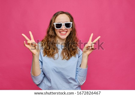 Stylish caucasian girl in a pale blue t-shirt and 8-bit glasses shows two fingers abstract peace smiling on a pink background. Close up.