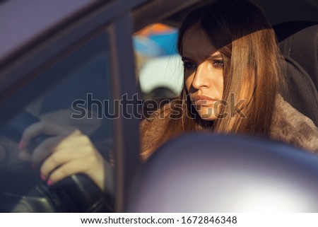 Portrait of young beautiful caucasian woman girl sitting in the car driving in sunny day wearing fur jacket coat looking to the road holding the wheel bored face traffic