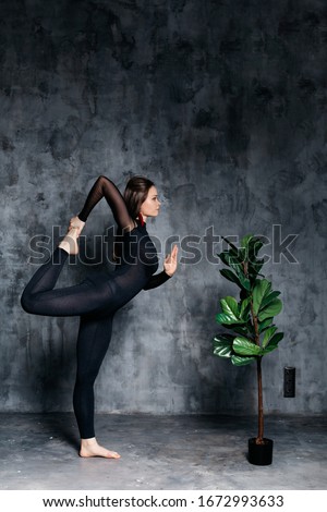 A photo of a beautiful brunette girl who is standing in a yoga pose on one leg, is holding her second leg with her hand. Nearby is a flower. Photo on a gray background