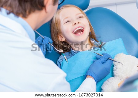 Little cute smiling girl is sitting in dental chair in clinic, office. Doctor is preparing for examination of child teeth with tools, amusing patient with bear toy. Visiting dentist with children. Royalty-Free Stock Photo #1673027014