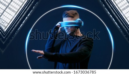 Guy using VR helmet scrolling invisible screen while interacting with virtual reality. Blue neon light. Augmented reality, future technology, game concept. Black minimalism.