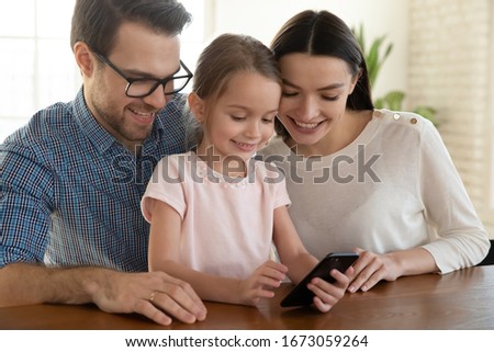 Happy family couple sitting with joyful small daughter at table, watching funny video together at home. Smiling little kid girl playing game on mobile phone with pleasant loving parents, head shot.